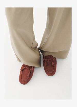 Osoba ubrana w beżowe spodnie o szerokich nogawkach i rdzawo-brązowe mokasyny zamszowe z chwostami na białej podłodze.