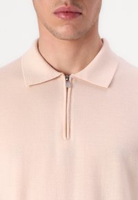 Polo tričko v světle růžové látce s žebrovaným límcem a zapínáním na polovinu zipu. Hladká textura s jemnými švy.