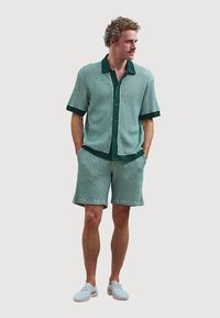 Kurzärmliges Strickoberteil und Shorts-Set in Türkis mit dunkelgr�ünen Akzenten. Verfügt über einen Button-Down-Kragen und strukturierten Stoff. Kombiniert mit hellen Schuhen.