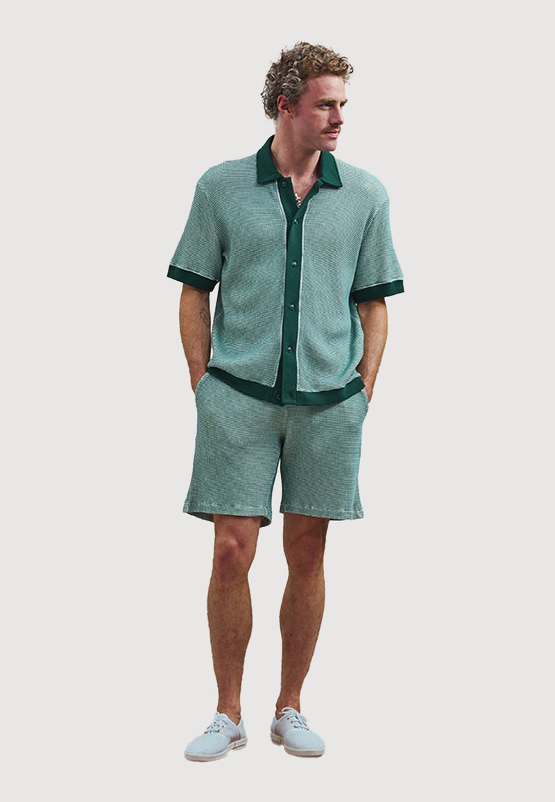 Kurzärmliges Strickoberteil und Shorts-Set in Türkis mit dunkelgrünen Akzenten. Verfügt über einen Button-Down-Kragen und strukturierten Stoff. Kombiniert mit hellen Schuhen.