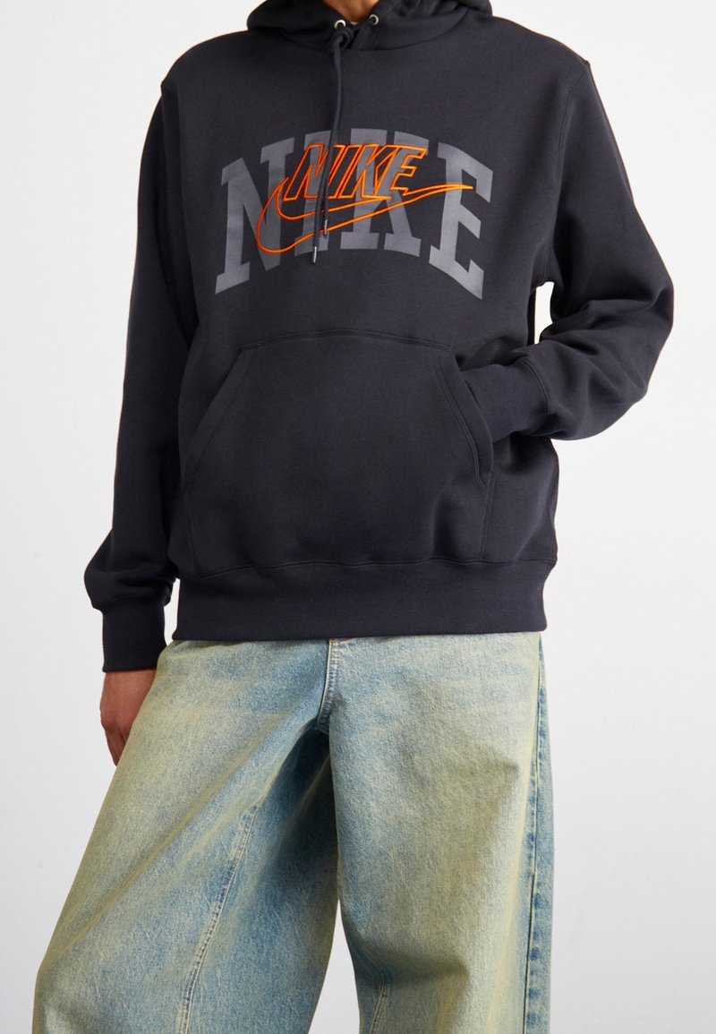 Svart huva sweatshirt med grå och orange Nike-logotyp. Har en framficka och justerbart snöre. Bärs med ljusblå oversized jeans.