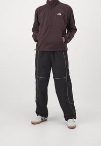Pullover in pile di colore viola scuro con zip a metà, abbinato a pantaloni neri con cuciture bianche e sneakers sportive bianche.