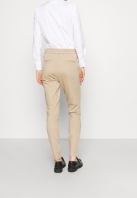 Person klädd i beige slim-fit byxor med elastisk midja och svarta skor, står mot en enfärgad vit bakgrund med ryggen vänd mot kameran.