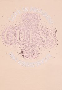 Lyserosa t-skjorte med rhinestein-dekorasjoner som danner ordet "GUESS" og buet tekst som leser "LOVE IN PROGRESS" og "ETABLERT 1981."