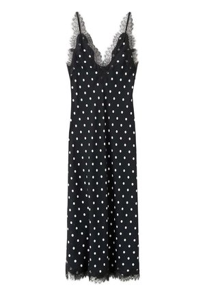 Femme aux longs cheveux marchant dos à nous, vêtue d'une robe midi noire à pois blancs et de sandales à talons noires dans une pièce blanche.