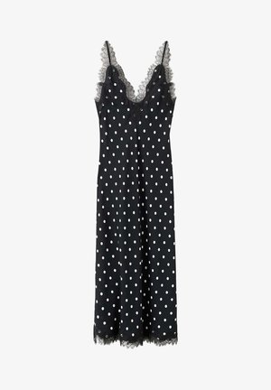 Femme aux longs cheveux marchant dos à nous, vêtue d'une robe midi noire à pois blancs et de sandales à talons noires dans une pièce blanche.