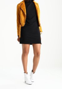 Veste de moto en cuir jaune sur une robe noire ; bottines blanches. La veste présente des fermetures éclair et des détails en bouton. Textures lisses.