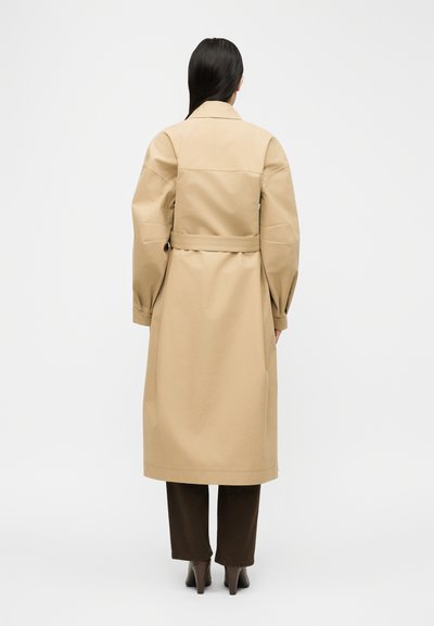 Άτομο με μακριά σκούρα μαλλιά, φοράει μπεζ παλτό με ζώνη, σκούρο παντελόνι και μποτάκια με τακούνι, στέκεται γυρισμένο πλάτη σε λευκό φόντο.