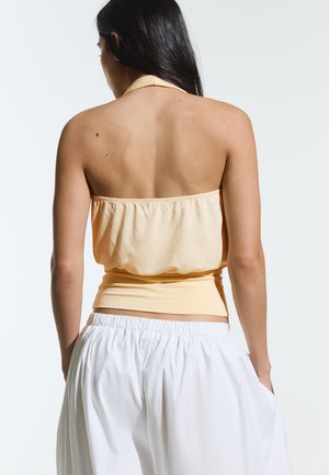 Frau mit langen dunklen Haaren, die ein helles Pfirsich-farbenes Neckholder-Top und lockere weiße Hosen trägt, von hinten vor einem schlichten hellen Hintergrund gezeigt.