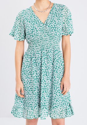 Femme portant une robe verte à motif floral blanc, manches courtes, col en V, taille smockée et ourlet à volants, accessoirisée d'un collier chaîne argenté.