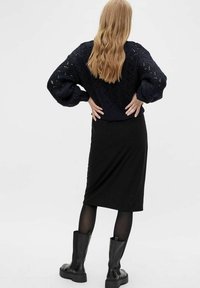 Kvinna med långt, vågigt blont hår, klädd i en marinblå strukturerad tröja, svart midi-kjol, genomskinliga svarta strumpbyxor och svarta kraftiga stövlar, står med händerna på höfterna.