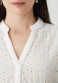 Hvit bluse med broderte hull, mandarin-krage og knappestolpe. Teksturert, lett stoff med små sirkulære utskjæringer.