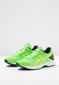 Zielone siatkowe buty biegowe z żółtymi akcentami, białą podeszwą środkową, czarną podeszwą zewnętrzną i płaskimi sznurowadłami. Posiadają wyściełany kołnierz i język.