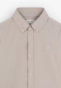 Camisa beige de botones hecha de algodón, con un cuello clásico, botones blancos y un pequeño emblema de calavera y huesos cruzados en azul claro.