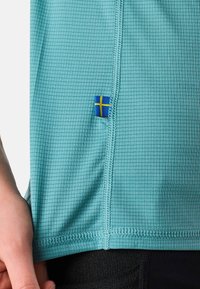 Nærbillede af turkis tekstureret stof med synlige syninger og et lille blåt og gult svensk flagmærke nær sømmen.