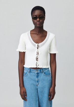 Top corto de canalé blanco con escote en V profundo y lazo en la parte frontal, con mangas cortas y fruncidas. Combinado con jeans de pernera ancha de color azul claro.