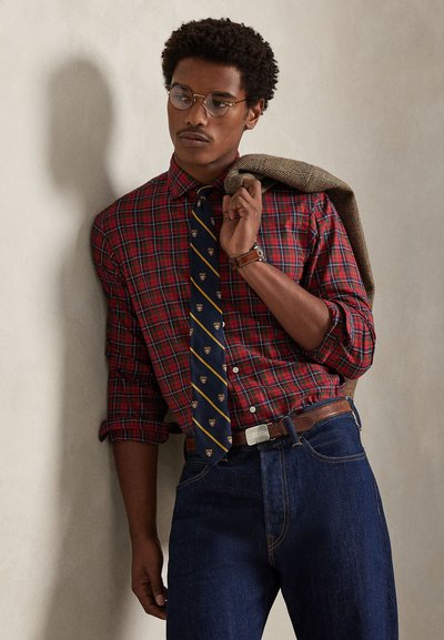 Czerwona i niebieska koszula w kratę zapinana na guziki z podwiniętymi rękawami, granatowy krawat z złotymi akcentami, dżinsy, brązowy skórzany pasek oraz brązowa marynarka o teksturowanej powierzchni.