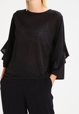 Femme portant un haut noir ample avec des manches à volants superposés et un pantalon noir, debout les mains dans les poches sur un fond blanc.