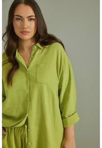 Camisa de gran tamaño de color verde lima con botones, con un bolsillo en el pecho, mangas enrolladas y textura suave. Presenta un ajuste casual y relajado.