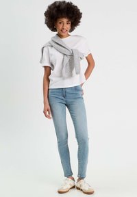 T-shirt blanc, jeans skinny bleu clair, pull gris drapé sur les épaules et baskets claires avec des accents beiges. Modèle posé contre un fond uni.