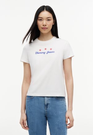 Donna con lunghi capelli scuri che indossa una t-shirt bianca "Tommy Jeans" con tre stelle rosse, abbinata a jeans di denim blu, in piedi contro uno sfondo bianco.