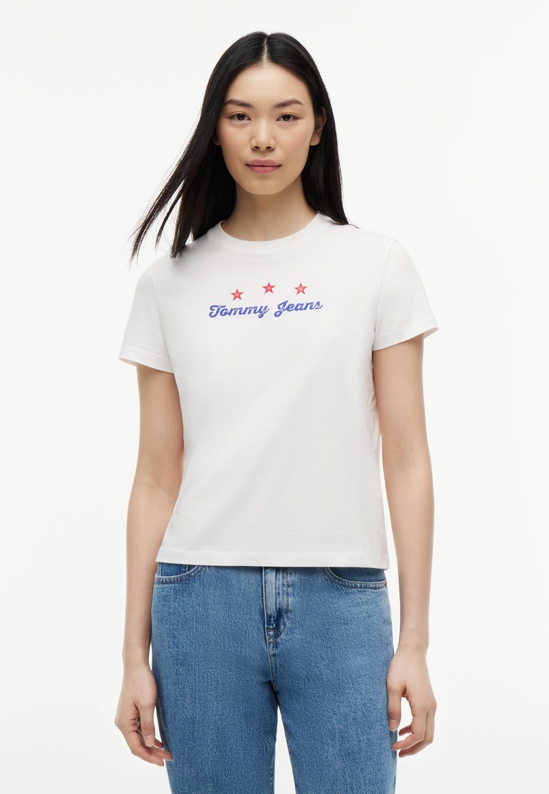 Femme aux longs cheveux foncés portant un t-shirt blanc "Tommy Jeans" avec trois étoiles rouges, associée à un jean en denim bleu, debout devant un fond blanc.