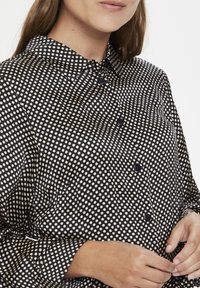 Svart skjorta med knappar och krämfärgade prickar, kragar, slät tyg och långa ärmar med texturerade manschetter.