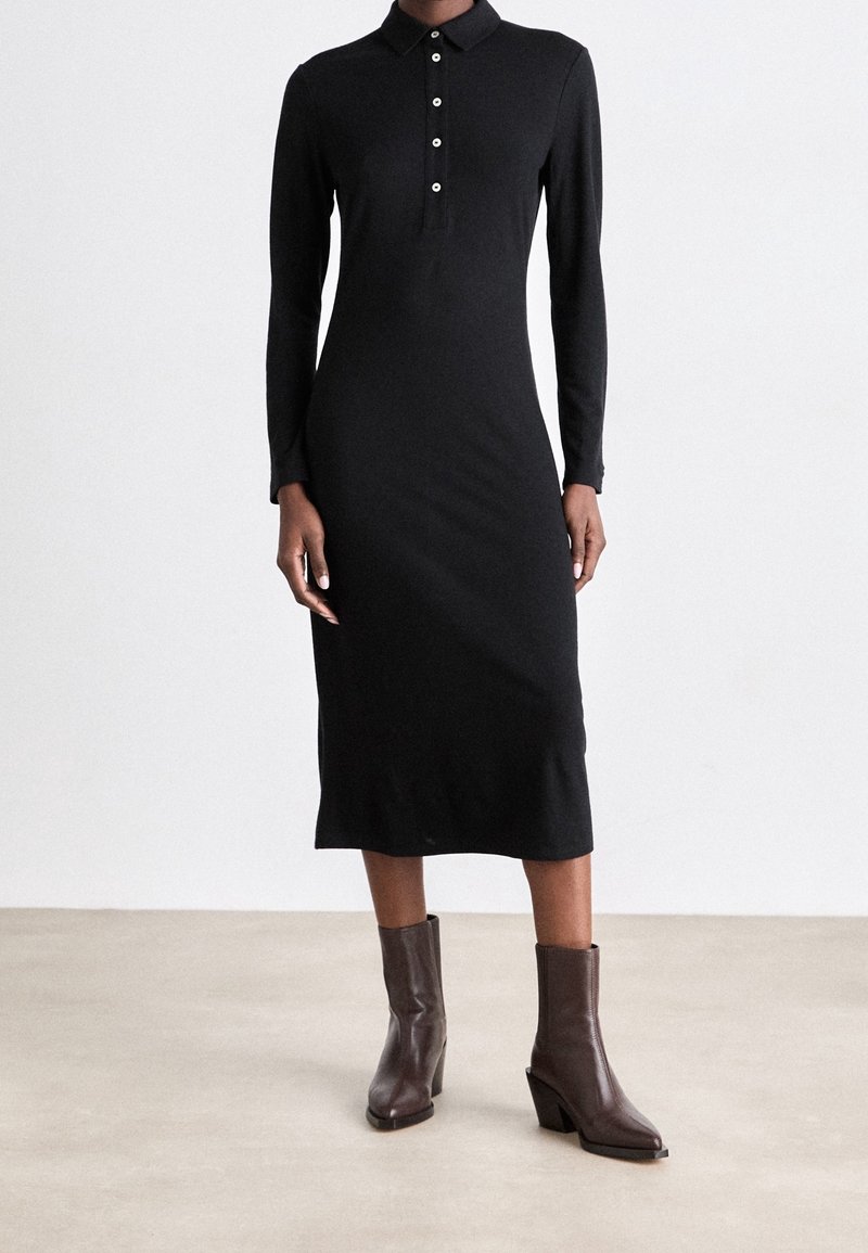 Robe noire à manches longues avec col et patte de boutonnage ; coupe ajustée, longueur genou, réalisée en tissu lisse. Bottines marron.