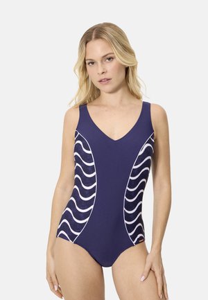 Mujer con un traje de baño de una pieza azul marino con franjas onduladas blancas en los costados, de pie contra un fondo claro y liso.