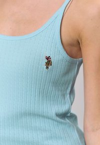 Šviesiai mėlynas ribuotas liemenėlės tipo marškinėlis su mažu siuvinėtu lopeliu, vaizduojančiu personažą. Turi plonas petnešėles ir aptemptą siluetą.