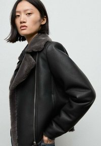 Giubbotto di pelle nera con collo in pelliccia grigia, dotato di chiusura con zip e vestibilità rilassata. Texture liscia e maniche oversized.