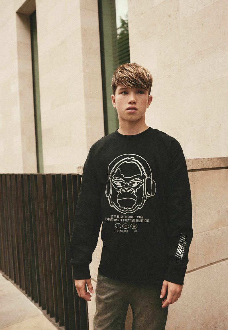 Adolescent garçon aux cheveux courts portant un sweat-shirt noir avec un visage de gorille et un texte, debout dehors près d’un mur en pierre et une rambarde en métal.