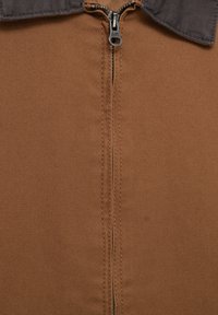 Brązowy materiał o gładkiej fakturze z centralnym zamkiem błyskawicznym i kontrastującym szarym kołnierzem. Szwy są widoczne wzdłuż krawędzi.