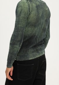 Grüner, gerippter Pullover mit langen Ärmeln und Farbverlauf, figurbetont geschnitten mit kontrastfarbigen Rippen am unteren Saum, kombiniert mit schwarzen Hosen.