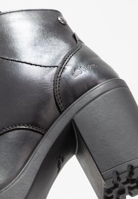 Bottine en cuir noir à finition brillante, design incurvé et talon épais. Présente des coutures et un détail de logo embossé sur le côté.