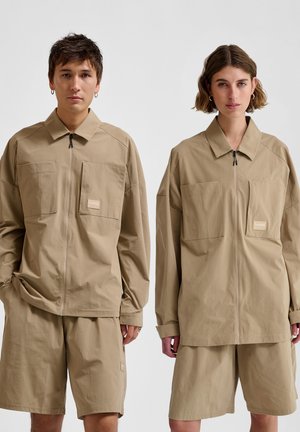 Beige oversized jack en shorts gemaakt van lichtgewicht stof. De jack heeft een ritssluiting aan de voorkant en grote zakken; de shorts zijn voorzien van zijzakken.
