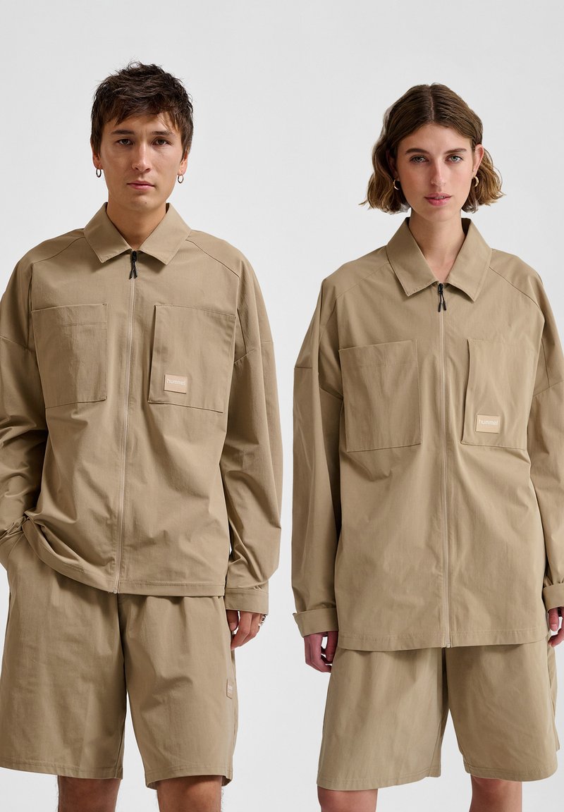Beige oversized jack en shorts gemaakt van lichtgewicht stof. De jack heeft een ritssluiting aan de voorkant en grote zakken; de shorts zijn voorzien van zijzakken.