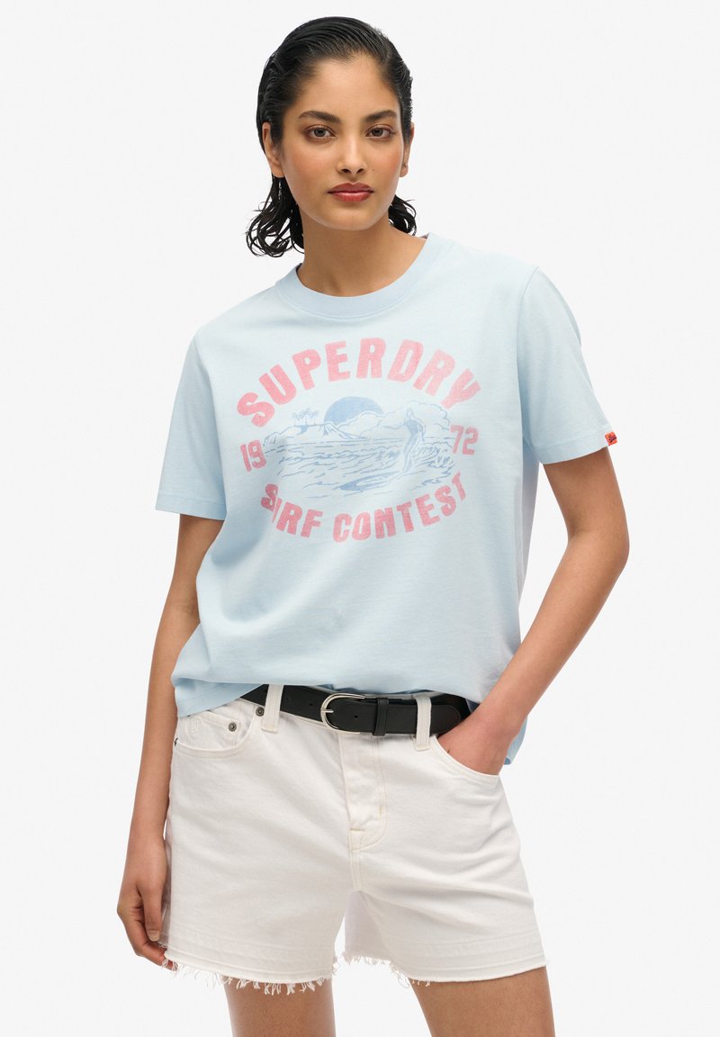 Camiseta de algodón azul claro con gráfico "SUPERDRY" en rojo y rosa, combinada con pantalones cortos de mezclilla blanca con dobladillos deshilachados y un cinturón negro.