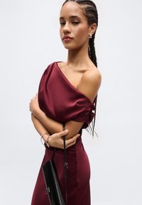 Kvinne med flettefrisyre iført en burgunderrød sateng kjole med én skulder, holder en svart clutch-veske og har på seg sølv armbånd og øredobber.