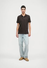 Σκούρο καφέ πόλο μπλουζάκι με γιακά και κουμπί, συνδυασμένο με ανοιχτό μπλε, κανονικής γραμμής τζιν και μπεζ αθλητικά παπούτσια. Απλός σχεδιασμός χωρίς ορατά μοτίβα.