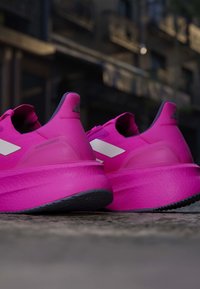Par lyserøde Adidas løbesko med tykke såler placeret udendørs på fortov mod sløret bygningsbaggrund.