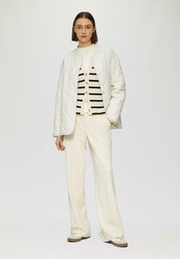 Gepolsterte weiße Jacke über einem schwarz-weißen gestreiften Cardigan, kombiniert mit cremefarbenen Schlaghosen und beigen Slippern. Einfaches Design, strukturierter Stoff.