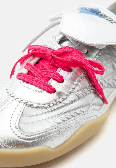 Chaussure de sport argentée avec des coutures texturées, des lacets rose vif noués en nœud et une semelle beige visible sur le bord inférieur.