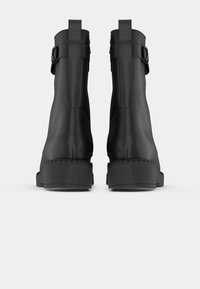 Bottes noires à la cheville avec une finition lisse et mate. Elles sont dotées d'une sangle avec des garnitures dorées, d'un talon carré et d'une semelle texturée.