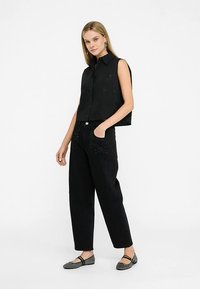 Zwart mouwloos crop topje met knoopsluiting, gecombineerd met zwarte hoge taille broek met voorzakken en subtiele versieringen.