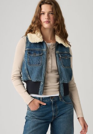 Junge Frau trägt eine Jeansweste mit Shearling-Kragen über einem beigen langärmeligen Henley-Shirt und blauen Jeans, Hand in der Tasche.