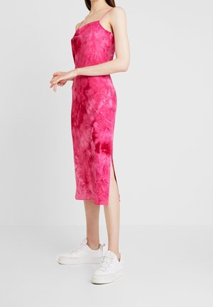 Woman wearing a sleeveless, pink tie-dye midi dress with a side slit and white sneakers, standing against a plain light background.