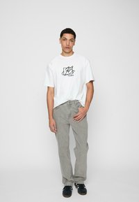 Witte t-shirt met korte mouwen met zwart grafisch ontwerp, gecombineerd met lichtgrijze, iets getailleerde broek en zwarte sneakers. Eenvoudige, casual look.