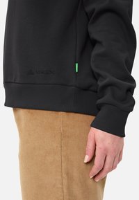 Felpa nera con polsini e orlo a costine, tessuto testurizzato, con un logo discreto in basso e un'etichetta verde sul lato.