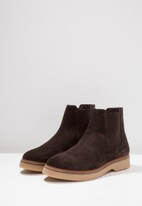 Botas Chelsea de ante marrón con paneles laterales elásticos y una suela de goma texturizada en tono marrón claro, con un detalle cosido alrededor de la base.